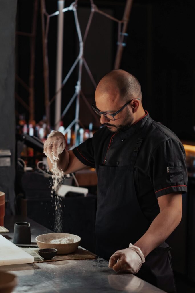 A focused chef sprinkles flour over a dish in a dimly lit restaurant kitchen.